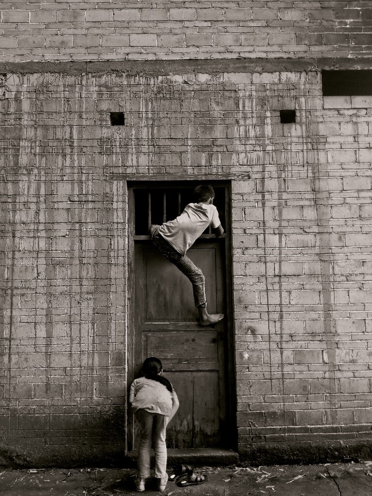 作品拍摄于广西壮族自治区贵港市平南县的一个小山村里,哥哥和弟弟出去玩的时候,忘带钥匙了,还好家里的这个后门是不用钥匙就可以打开的,两个孩子在想办法开门,哥哥爬上门拿棍子尝试伸进去打开门,好奇的妹妹透过门缝想要观察观察里面的情况。<br />
这个摄影作品记录的是广西山村里留守儿童生活的一个瞬间,我是一个农村留守儿童的过来人,在我们看来,其实在大山里农村的生活是非常自由自在的,哪里都是我们的玩乐之地。这个作品虽然只是一个生活瞬间的记录,但是充满了童真的气息,表现了大山里孩子的调皮和童年生活的趣味。