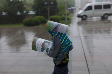 2014年4月26日，山东寿光蔬菜博览会，雨中一个孩子捡来宣传册挡雨，一本护着头，一本护着双手。