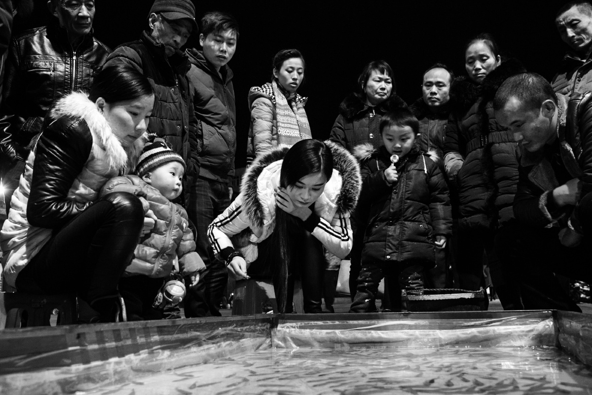 一群人正围在一起,看一个美女玩钓鱼游戏,美女专注的表情和围观的人