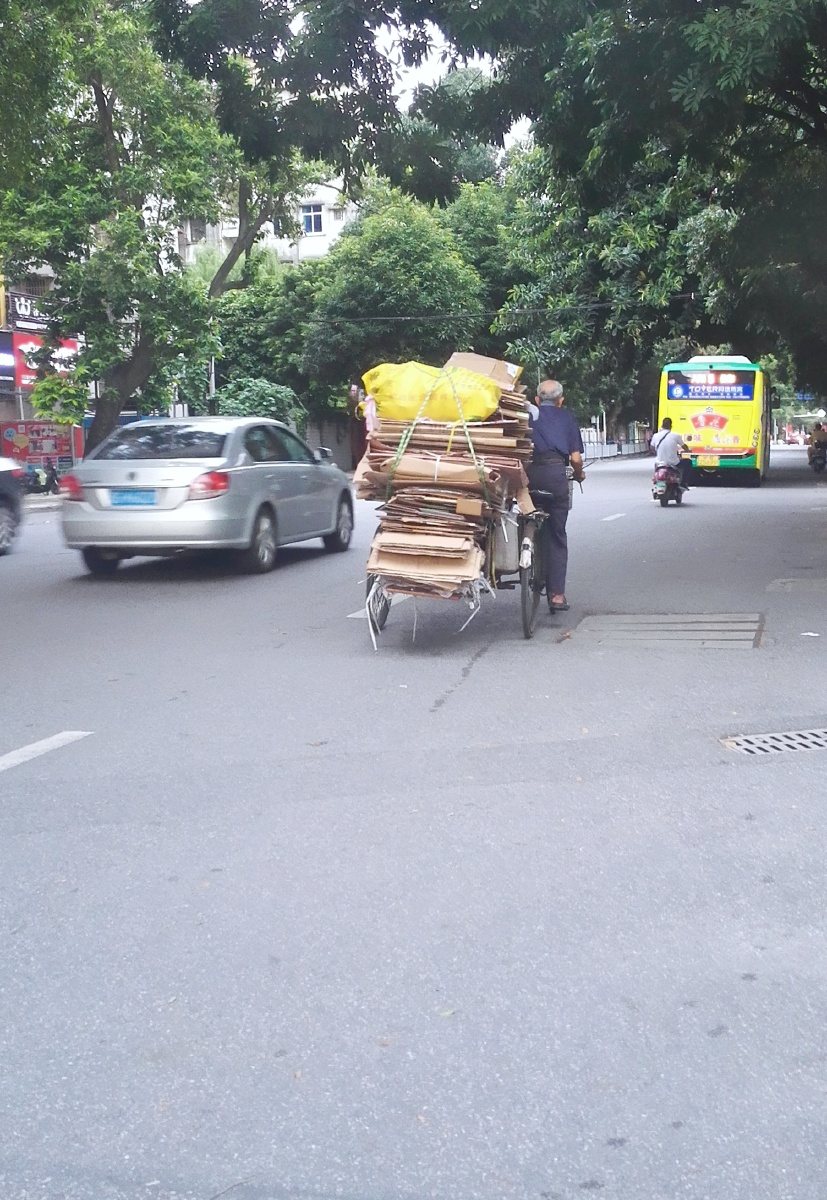 在繁忙的道路上,一位收废旧的老大爷吃力的踩着装满废纸皮人力三轮