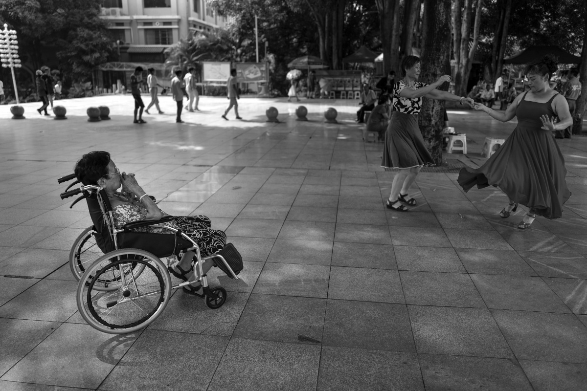在公园里一位卒中后遗症者坐在轮椅观看市民跳广场舞.2018年7月,百色