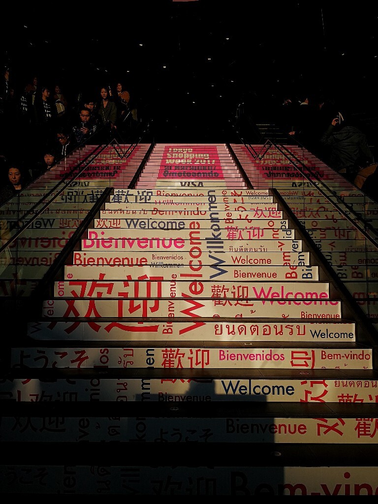 人们乘坐扶梯，让出被阳光打到的印有各国文字<br />
“欢迎”的阶梯。