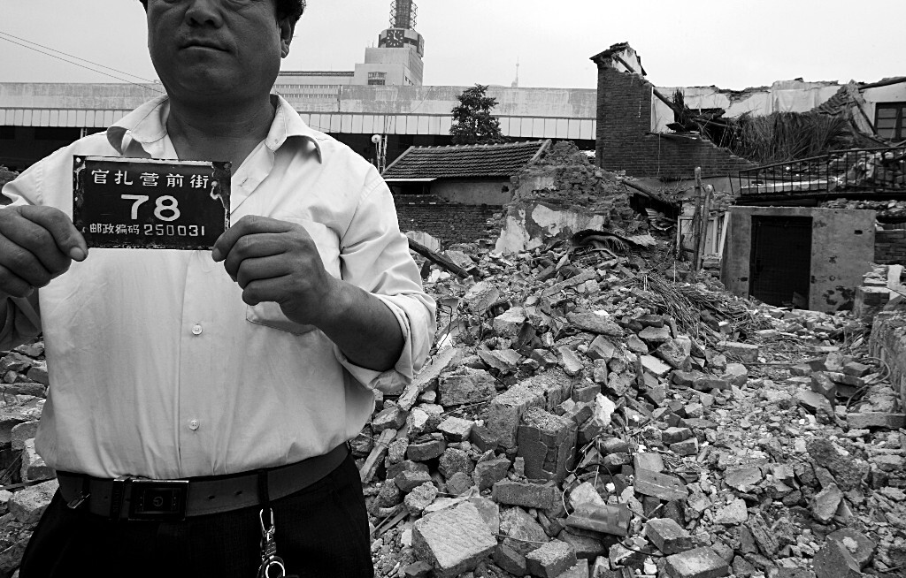 2012年8月26日，一位居民在拆除的老屋前，拿着自家的门牌号留影纪念。
