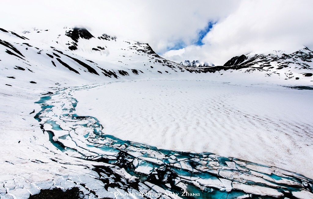 《冰河世纪》<br />
6月的瑞士，还有很多地方还是大雪封山。我们则开车穿行于夏季才开放的S弯山路之间，寻找可以拍摄的机位。这个湖离公路还有大半个小时，路上还被各种雪崩下来的积雪阻拦着。为了探个究竟，我们徒步前往，没想到冰湖刚开始消融，厚厚的冰层撕裂开来，恍如冰河世纪。当然更神奇的是遇到了一位素衣大妈，一个人在乱石堆中穿越雪山。我们带上了冰抓手杖，她则是运动鞋徒手，高手就在民间。