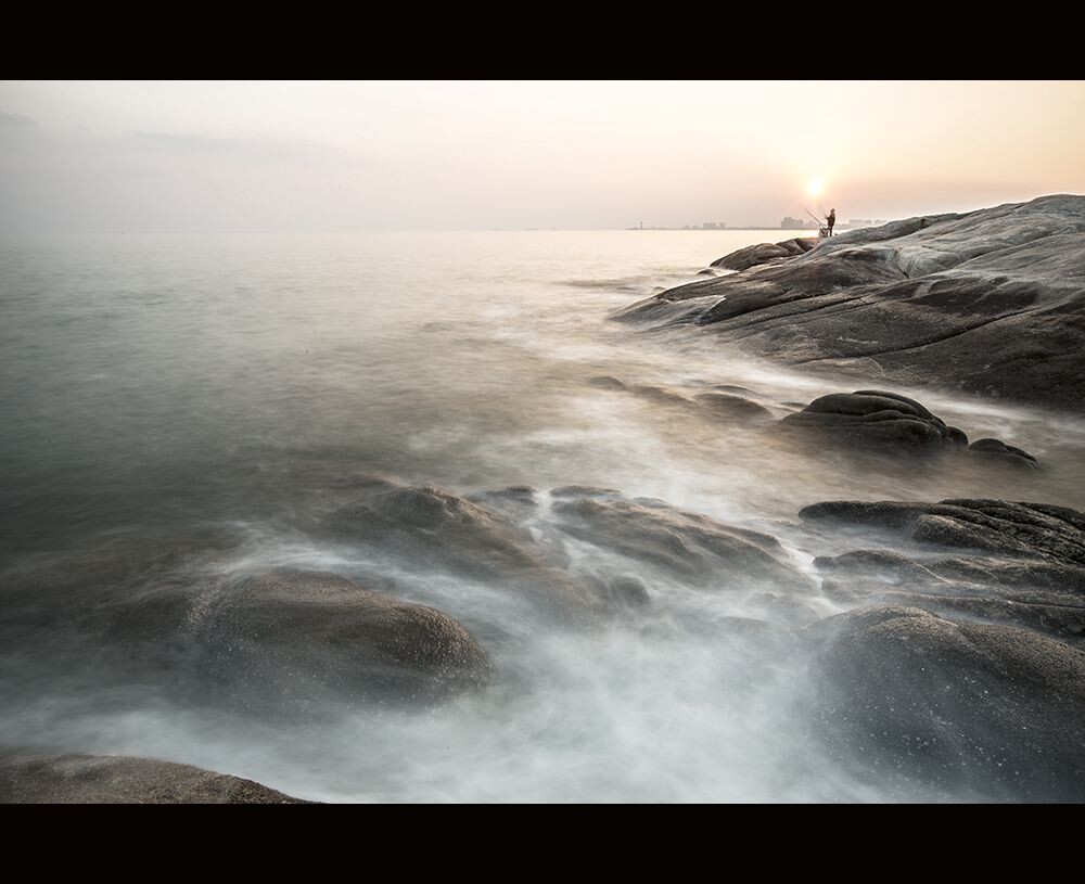 夕阳的暖色洒在海面上，海浪轻抚着礁石，垂钓者静静地站在风中，孤独地享受着独钓的快乐。。。