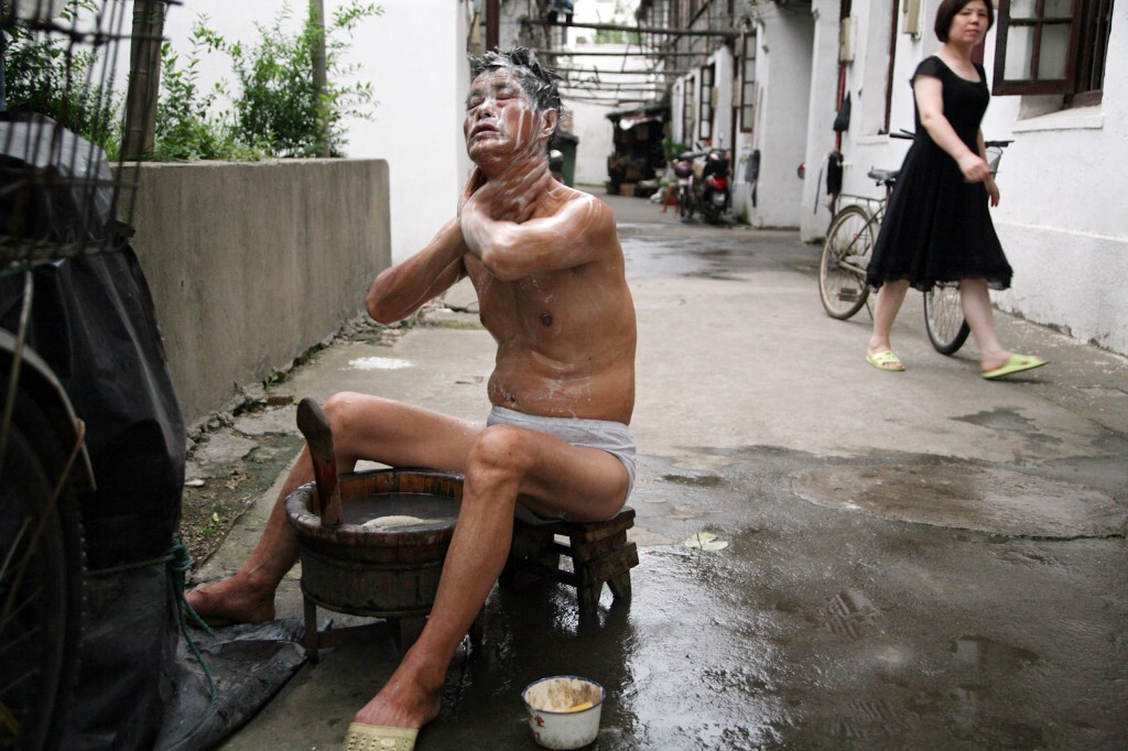 2010年6月30日,无锡盛新里,炎炎夏日,一位老人光着身子在弄堂里洗澡,一位妇女从他身后走过。老人用的木浴盆和装肥皂的搪瓷盆现已很少见。
