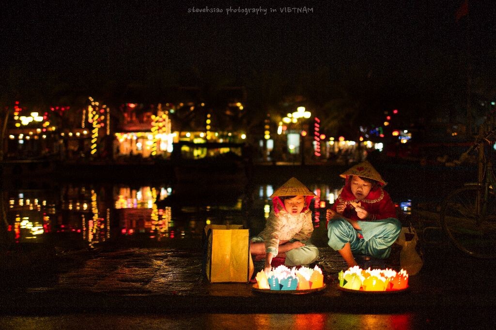Hoi An, Vietnam 会安古镇夜幕降临，在河边当地人开始向游客兜售纸船灯。和其他小贩揪着游客不放不同，两姐妹坐在河边悠闲地聊天，一幅纸船卖不卖得出去都无所谓的神态。