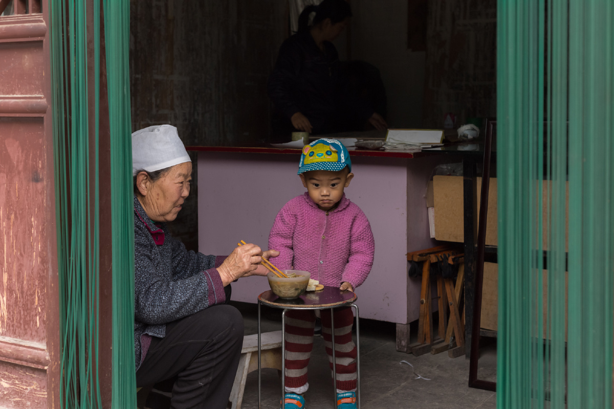 沿街店铺的一家人，到吃中午饭了，老人只有先喂孙子吃完，才会自己吃。