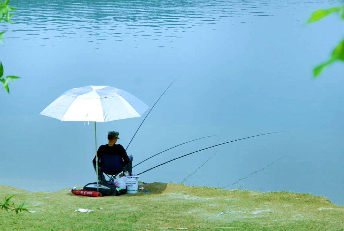 一位钓者悠闲地坐在湖边太阳伞下,享受钓鱼时光.