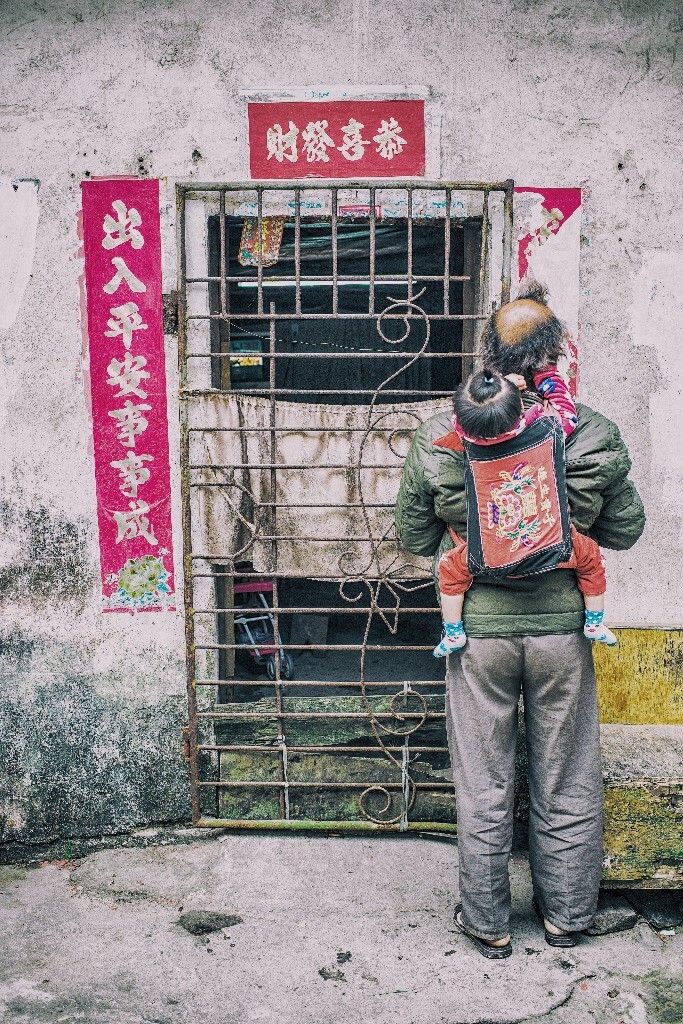 每年春节都去拍点应景的照片是我的习惯了，当又一次走进老城区，除了看到那些熟悉的年味，还偶遇了一些凄凉。路过这家门口时我没有做任何停留，我也看不到正在撕对联男子的正面，只是他的装束和背上扎着头发的孩子让这个年看起来过得并不富足。希望他们一家能过上好年好日子。The annual Spring Festival to take photos of the occasion is my habit, when once again into the old city, in addition to see those familiar flavor, also encounter some sad. I didn't do any stopping at the gate, and I couldn't see the face of the man who was tearing the couplets, but his clothes and the children with his hair on his back made it look like he was not rich this year. I hope the family will live a good life.