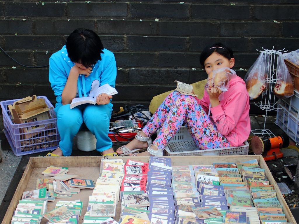 碑林博物馆旁边的马路上摆满了卖书画的摊子，有两个小朋友在卖小人书。不过她俩只顾着自己看书，完全不招呼顾客，在一旁摆摊子的家长呵斥了几声：别看了！