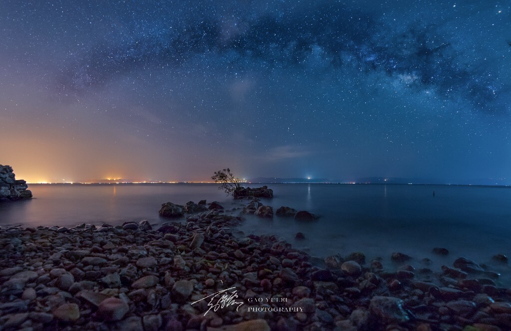 银河升起来，黑夜顺着湖岸边走，来到一个布满鹅卵石的浅滩，浅滩延伸至湖中，尽头竟然是一个小树生长在石间，孤寂而顽强。抬头一看，银河拱桥横跨湖面，远处的光污染正好给整个冷色的夜晚增添了几许暖调。壮美与孤寂，冷与暖，苍穹下的生命。