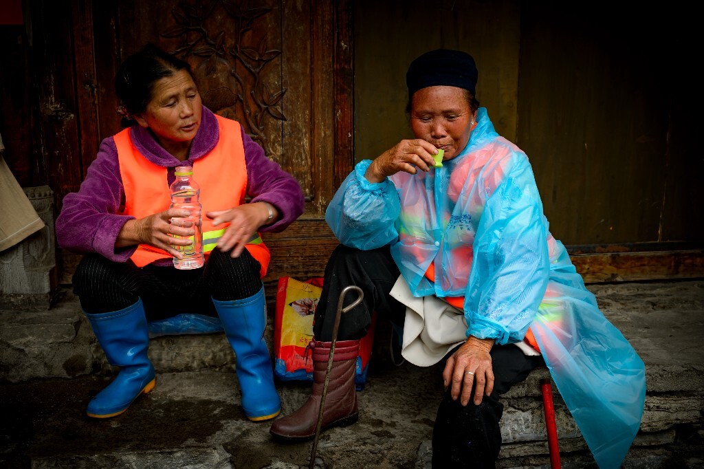 两位保洁员在屋檐下雨歇，汉族妹妹给苗族姐姐递上一满杯纯净水，又怕她喝得太急太快。