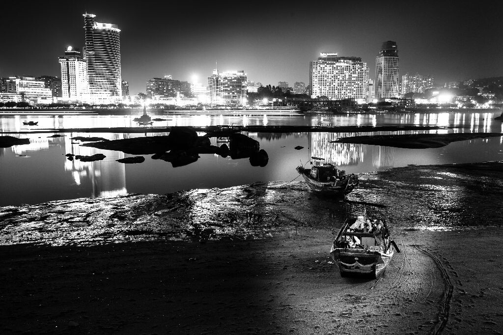 站在鼓浪屿拍摄对面的轮渡，潮水褪去，宁静的夜晚中，沙滩上停泊的船吸引了我，以海为生，以船家的人在船舱中休息。宁静的船只与喧闹都市形成了对比。PS：本来前面这船的夹板上有一个人在作业，可是架好脚架后，他已回了船舱。还是希望有人在画面中，会更生动。